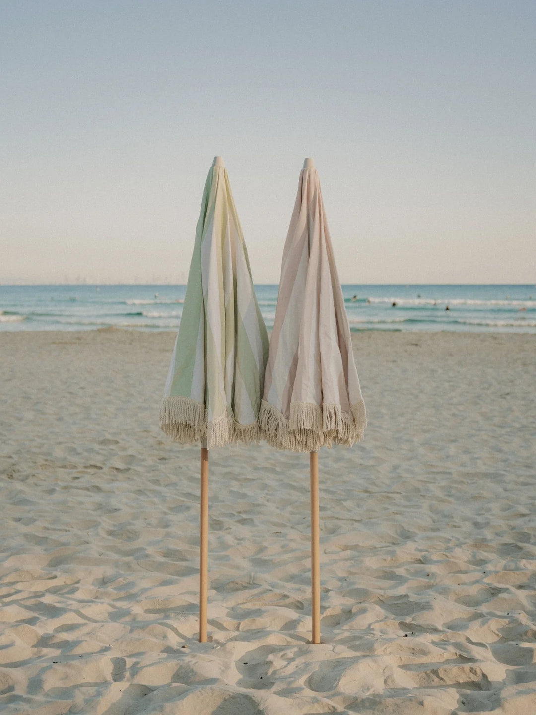 Salted Sage and Sand Drift beach umbrellas from House of Dune folded side by side on the sand near the ocean.