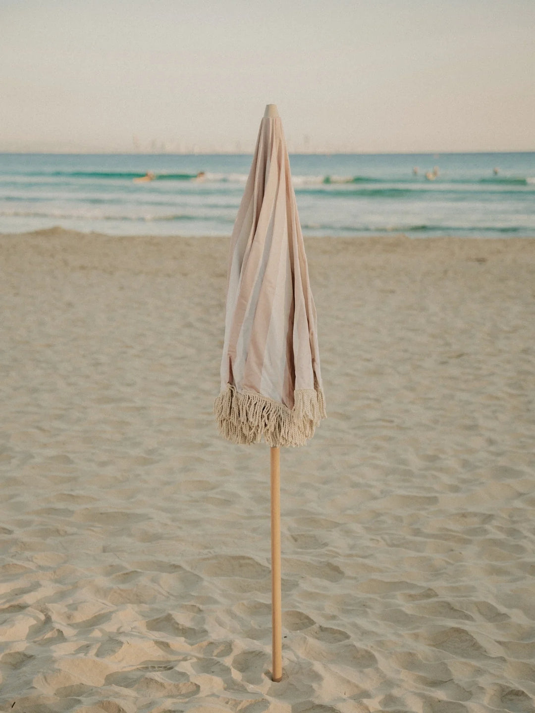 Sand Drift beach umbrella folded upright in the sand with ocean waves in the background, by House of Dune.