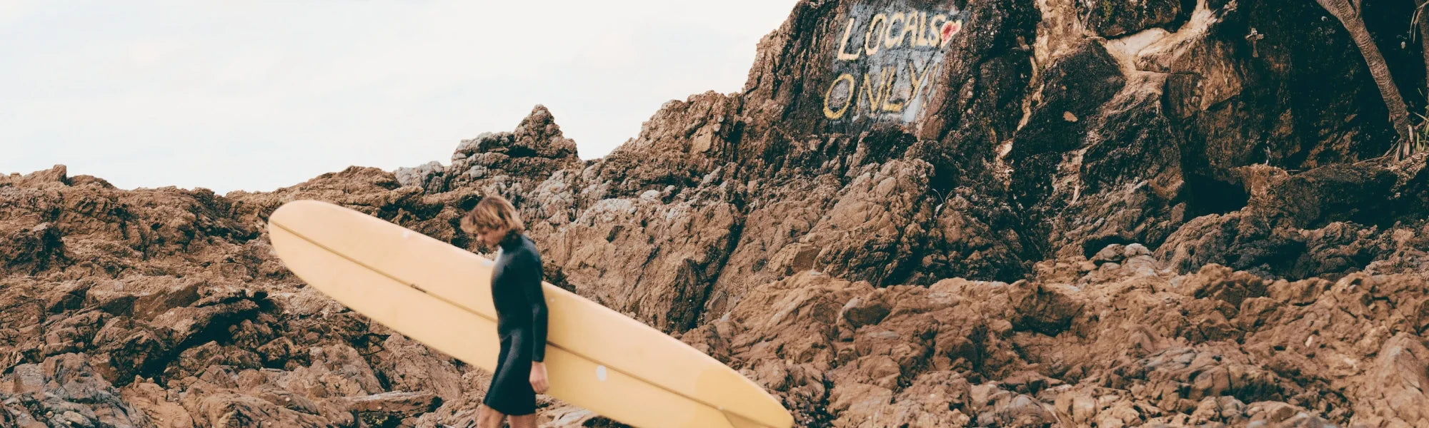 A surfer carrying a yellow longboard walks across rocky terrain in Byron Bay, with “Locals Only” painted on the cliffs in the background. The scene captures the laid-back coastal surf culture of Australia.