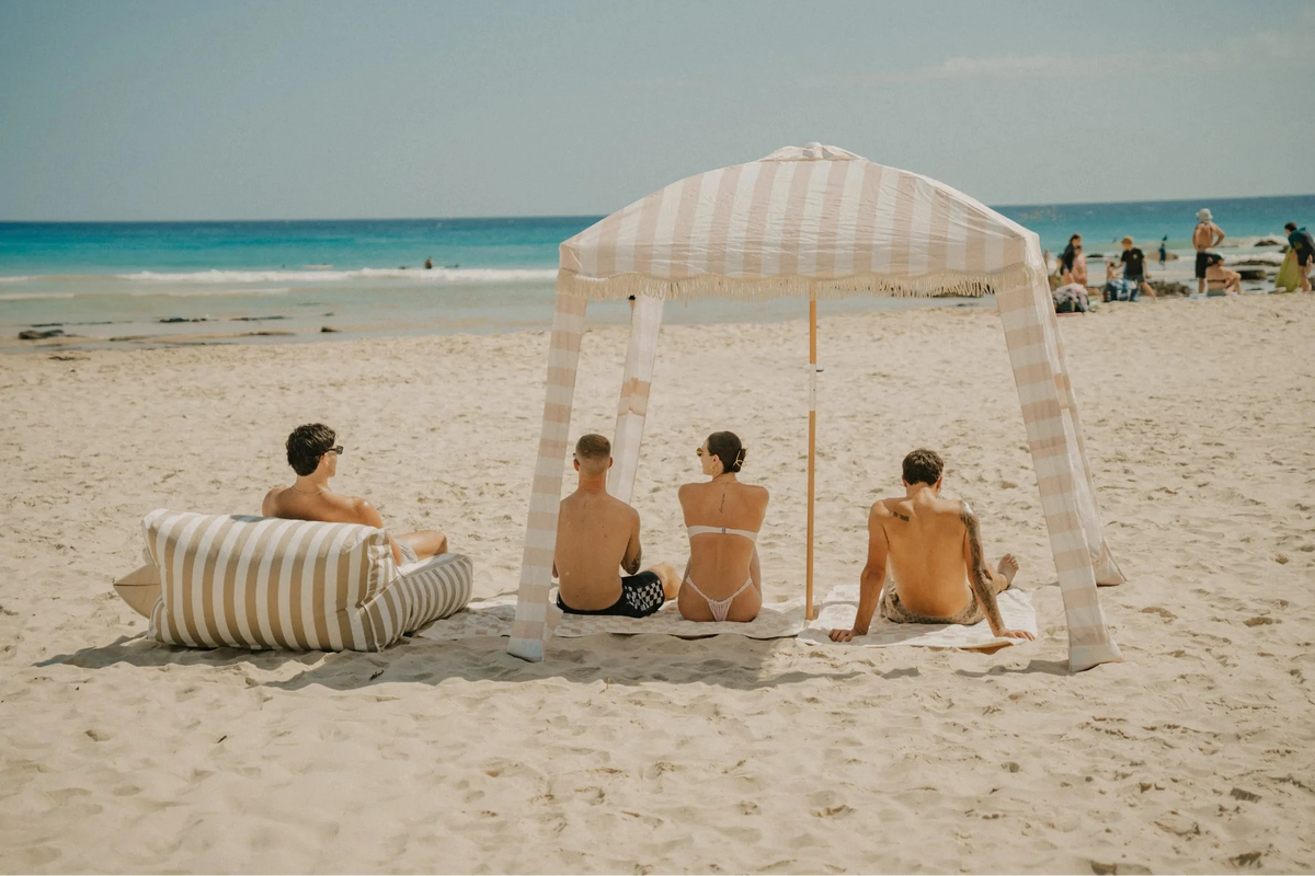 Group of four friends lounging under a beige and white striped House of Dune Sand Drift Cabana on the beach with matching loungers and towels, creating an elegant coastal shade setup.