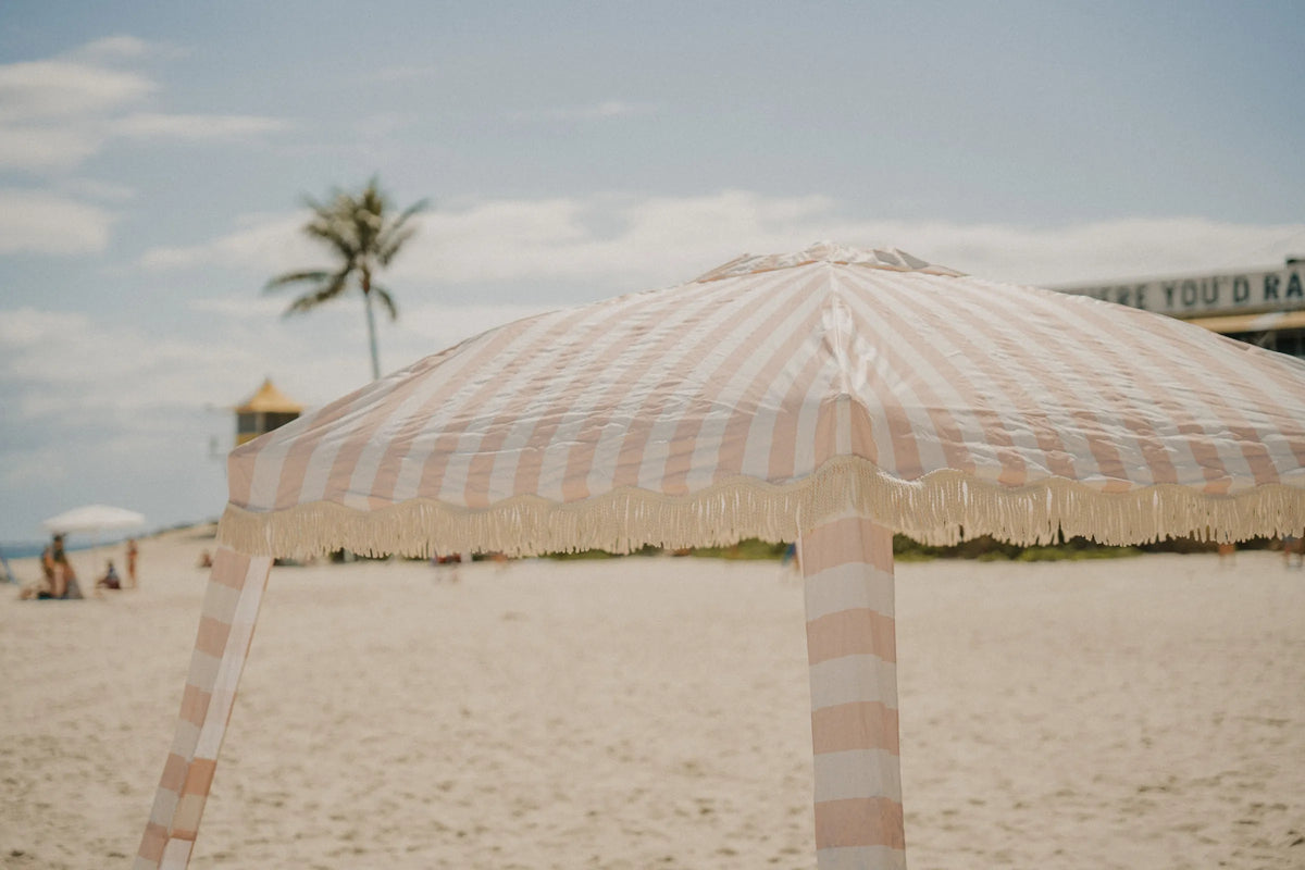 Close-up of the House of Dune Sand Drift Cabana in warm beige and white stripes with tassel fringe detailing on a sunny beach, showcasing a premium coastal shade canopy.