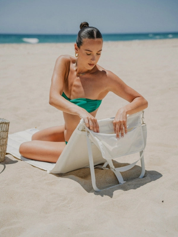 House of Dune Oyster Tide beach lounger made from recycled RPET, shown on a sandy beach with a woman sitting comfortably and accessing the integrated storage pocket.