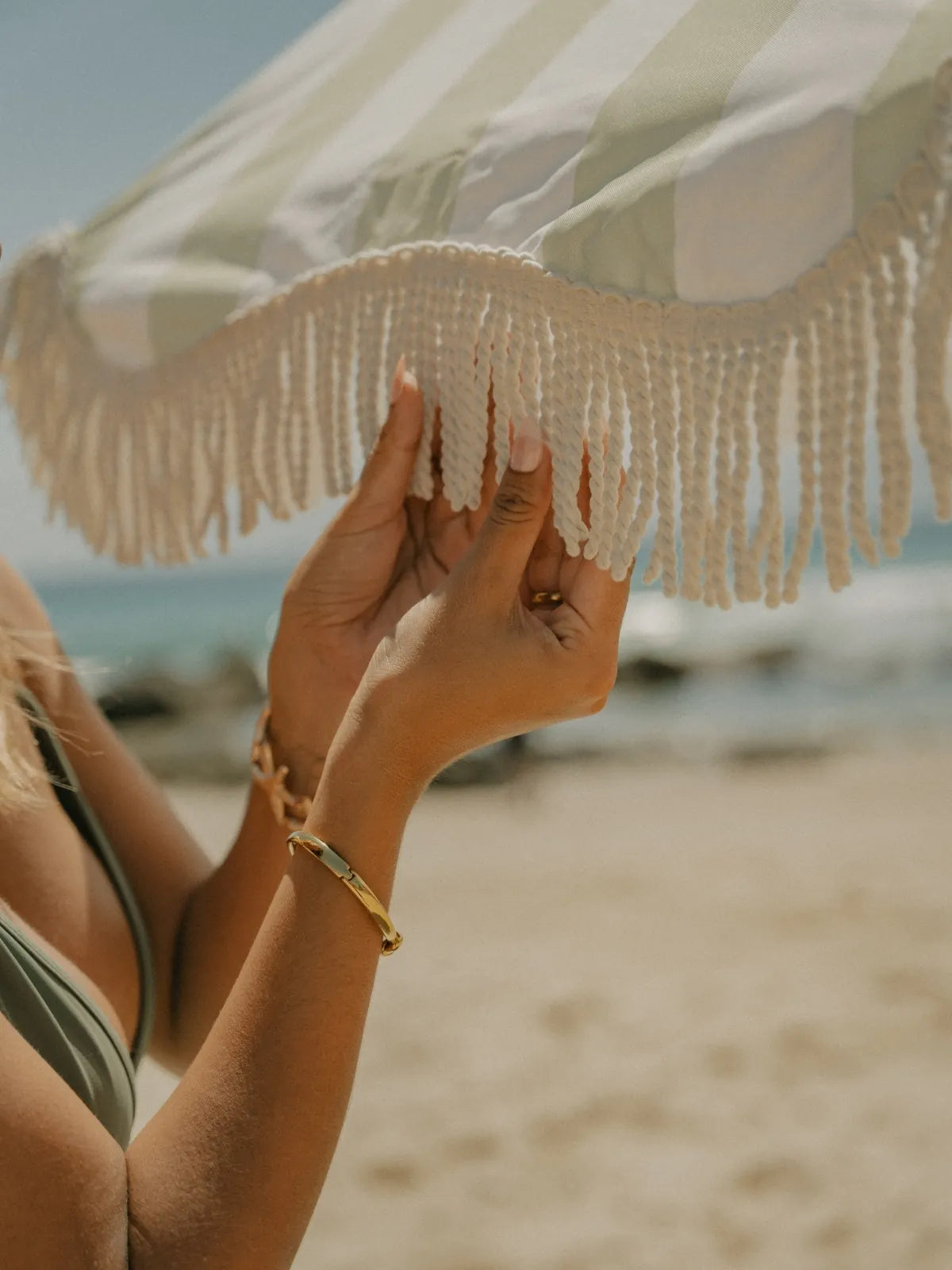 Hands adjusting the cream fringe of the Salted Sage beach umbrella by House of Dune on a sunny seaside day.