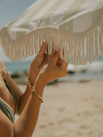 Hands adjusting the cream fringe of the Salted Sage beach umbrella by House of Dune on a sunny seaside day.