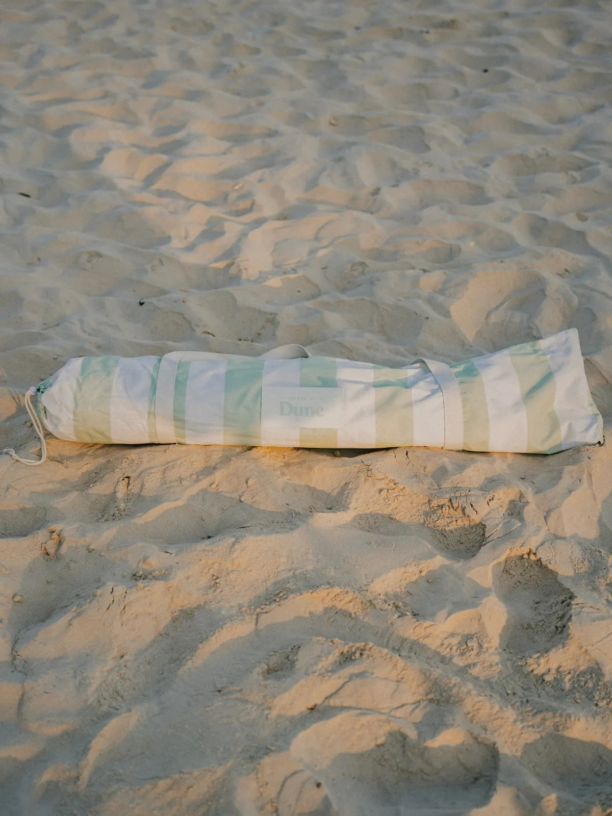 Salted Sage beach umbrella neatly packed inside its striped House of Dune carry bag resting on sand