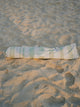 Salted Sage beach umbrella neatly packed inside its striped House of Dune carry bag resting on sand