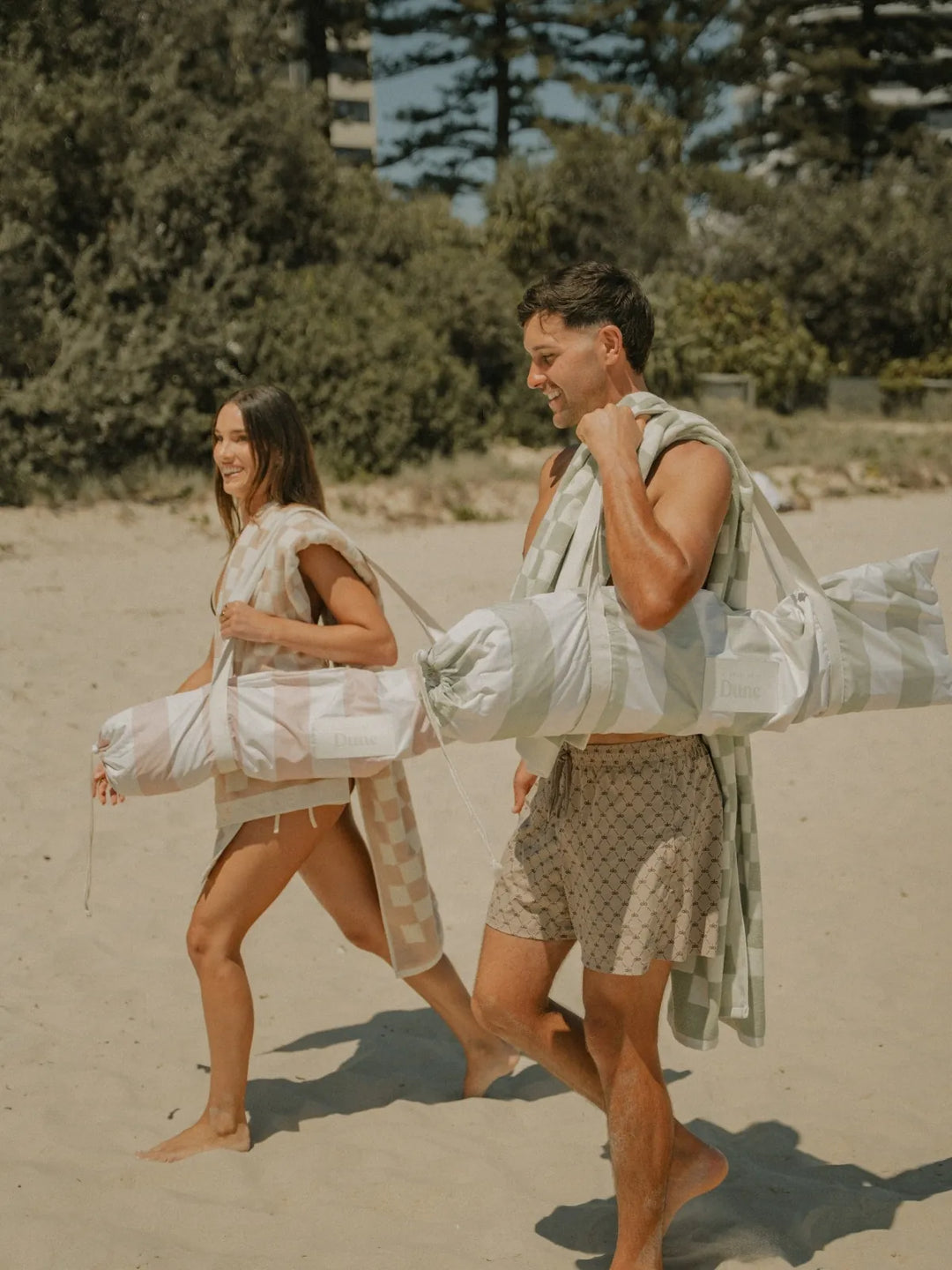 Two people walking along the beach carrying folded Salted Sage beach umbrellas from House of Dune.