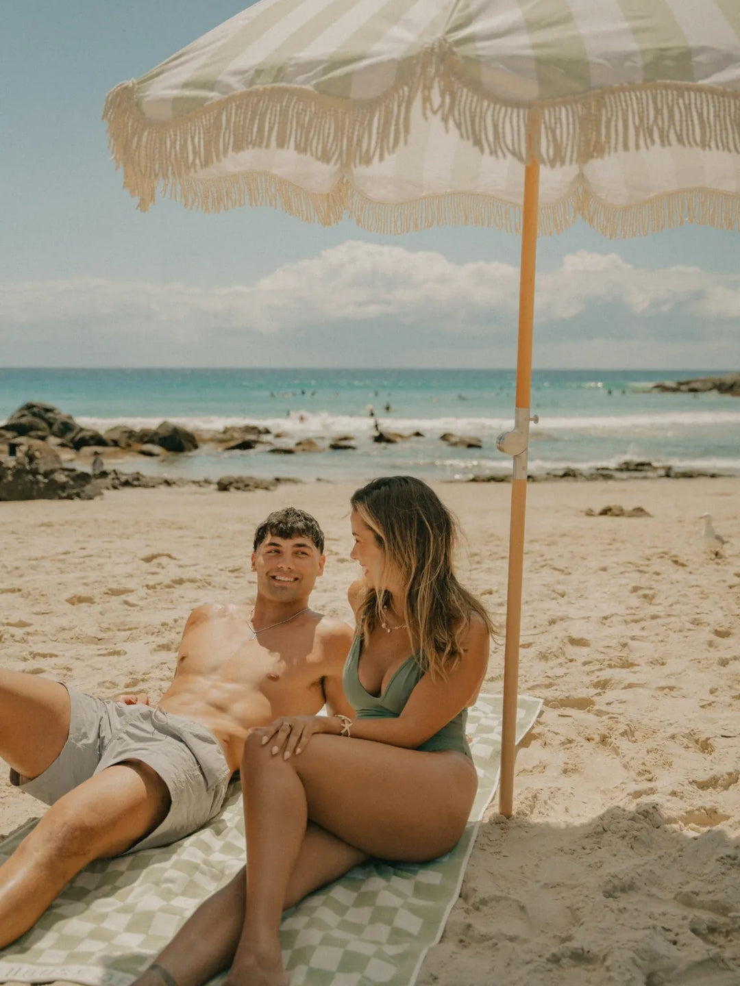 Couple relaxing together under the open Salted Sage beach umbrella by House of Dune with ocean waves behind them.