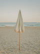 Salted Sage striped beach umbrella folded upright in the sand with calm ocean backdrop, by House of Dune.