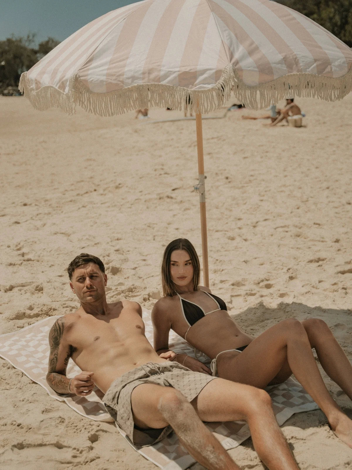 Man and woman lounging beneath the Sand Drift beach umbrella by House of Dune on a sunny coastal day.