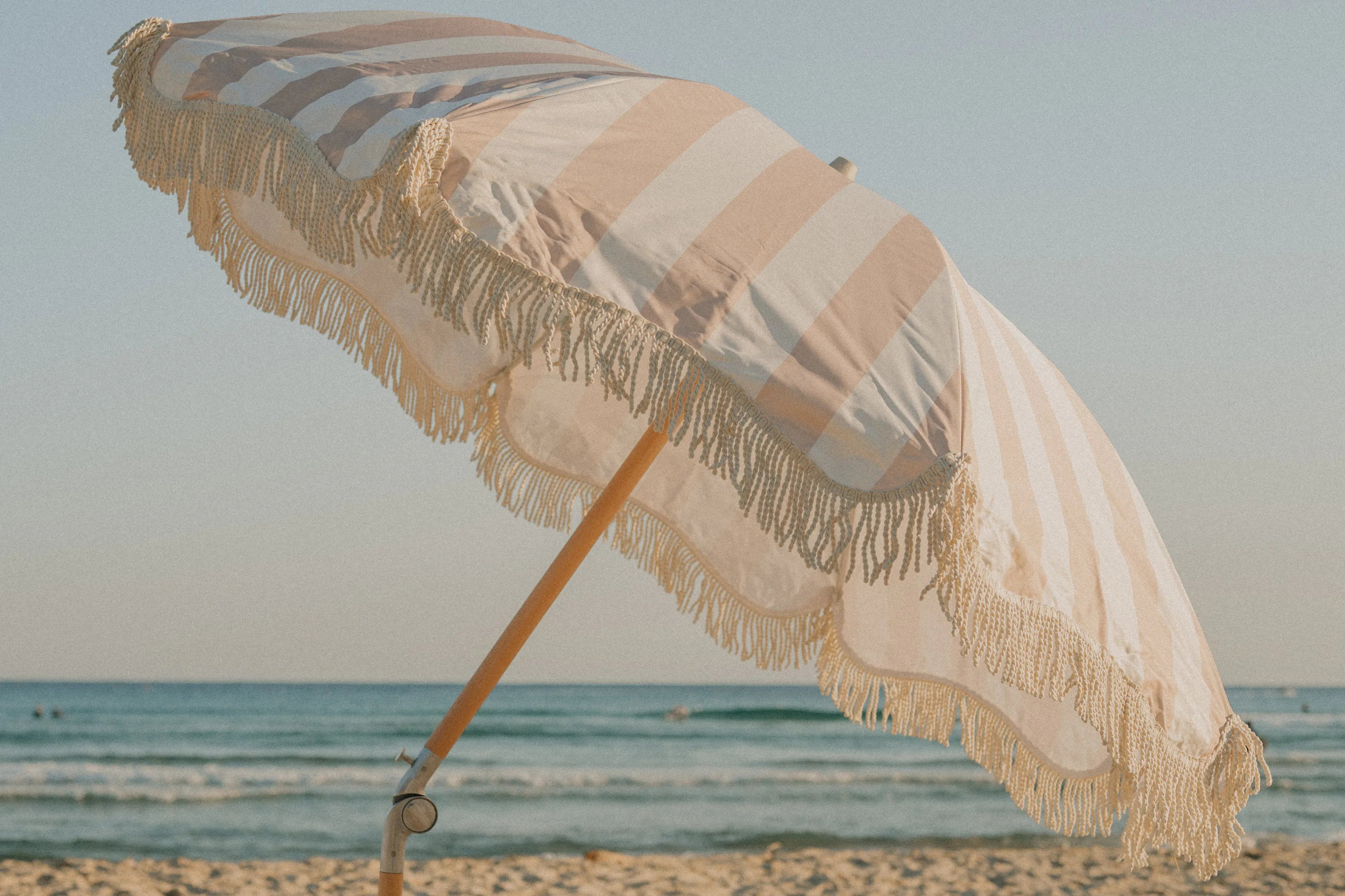 A Sand Drift fringed beach umbrella tilted over the sand by the sea