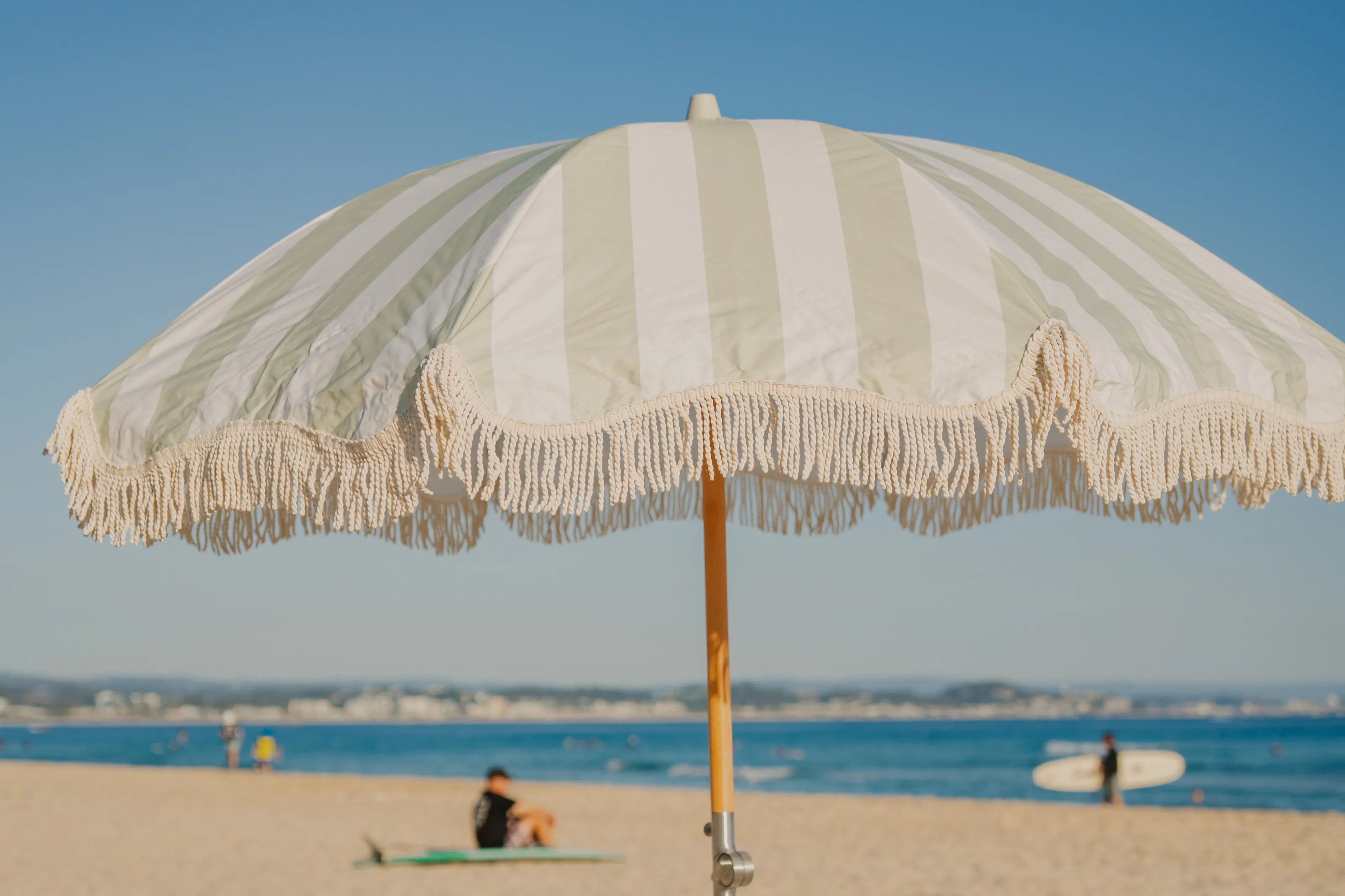 A Salted Sage fringed umbrella over a Salted Sage checked towel and a picnic basket on the sand