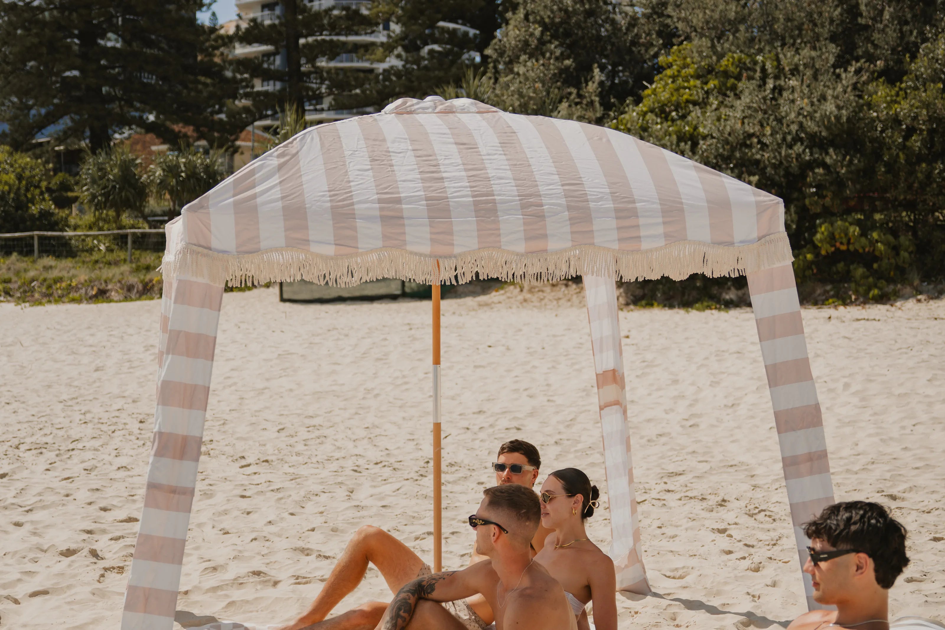 A Sand Drift striped beach cabana on the sand, with a floating lounger and friends in its shade