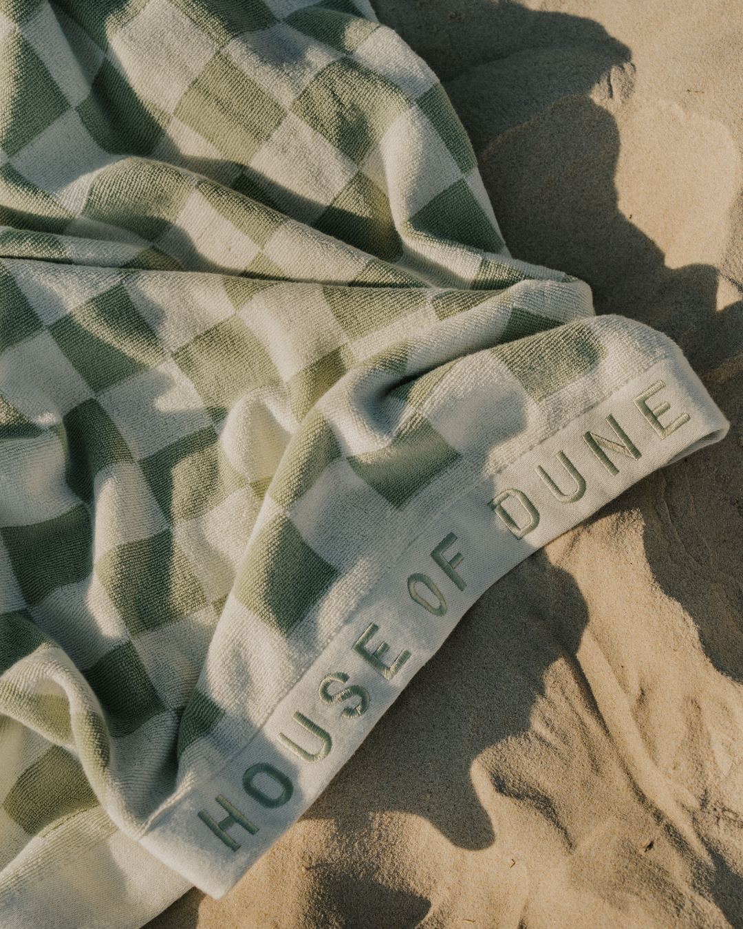 A Salted Sage Beach Towel with its woven House of Dune edge, lying on the sand